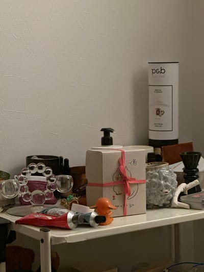 An overhead-lit, static interior scene in Suginami, Japan, displays a cluttered white shelf against a textured off-white wall. On the left, a transparent glass decorative object composed of interconnected spheres is positioned next to a dark cylindrical container and a smaller dark bottle with a dropper. In the immediate foreground, two squeezed tubes, one red and one light blue/grey, rest horizontally beside a small orange plastic figurine. Central on the shelf is a light brown cardboard box, possibly a soap or lotion dispenser, featuring a black pump top, a pink ribbon tied around it, and a simple illustration of a cat and a bee. To the right of the box, a grey, textured ceramic pot with a stylized face-like design is visible, adjacent to a piece of white coral and a dark, chalice-shaped decorative item. A tall, white cylindrical container stands on the far right, bearing the "p&b" logo, "GRAND VIN DE BORDEAUX," "1926 RENAISSANCE," and a coat of arms, suggesting wine packaging. A brown, possibly leather, object is partially visible behind the ceramic pot. The shelf's left support post is visible, and below the shelf, a portion of a reddish-brown, soft object is discernible. No people are present.