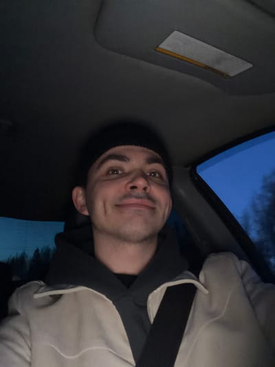 A man wearing a black beanie, a dark hooded shirt, and a light-colored jacket with a seatbelt across his chest is in the driver's seat of a car. The interior of the car is visible, including the sun visor. The sky is visible through the car window. No information exists to suggest the location in the city of Shaberdinka, Russia.