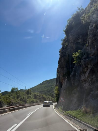 The image depicts a highway in Kruševica, Montenegro, under a clear blue sky with bright sunlight. A winding road with white lane markings is visible, with two cars traveling ahead. To the right, a steep, rocky cliff face is protected by a mesh netting system. Vegetation, including trees and shrubs, grows on the cliff and the surrounding hillsides. A railway track runs parallel to the road on the left. A triangular warning sign with a slippery road symbol is posted near the cliff.