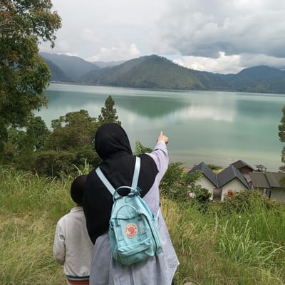 The image shows a woman wearing a hijab and a striped shirt, with a light blue backpack, pointing towards a scenic vista. Beside her, a child wearing a white jacket is standing. They are on a grassy hillside overlooking a large, calm body of water with lush green mountains in the background under a cloudy sky. Several houses with dark roofs are visible on the slope leading down to the water. The scene suggests a tourist or recreational area, possibly near Kalalengkio, Indonesia, given the presence of natural beauty and lakeside dwellings. The woman's gesture and the child's presence indicate they are observing or discussing the landscape. The overall atmosphere is serene and natural.