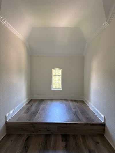 The image shows an empty, finished room with a raised platform floor covered in wood-look laminate flooring. The walls are painted a light cream color with textured wallpaper, and there is white crown molding along the top where the walls meet the sloped ceiling. A small, arched window with multiple panes is centered on the back wall, admitting natural light. The floor on the lower level also features the same wood-look laminate flooring. The scene is a generic interior view, likely of a residential property, but no specific Houston location is identifiable.