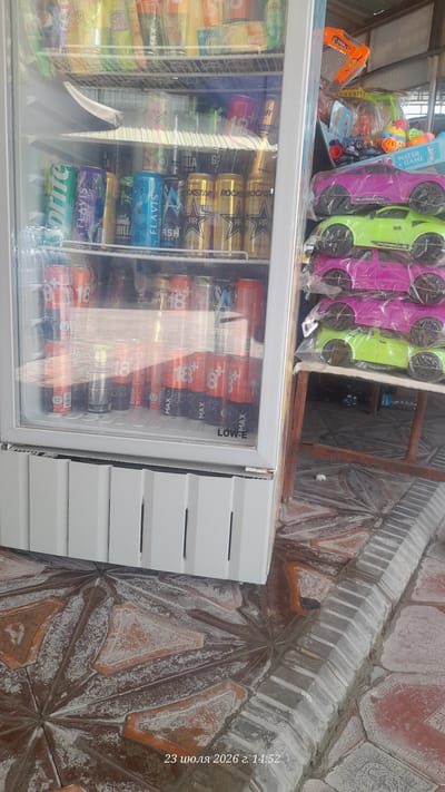 This is a frontal medium shot of a refrigerator filled with various drinks, primarily energy drinks and sodas, in a retail setting. The refrigerator has a glass door, revealing multiple shelves stocked with cans. Prominently visible are cans with branding such as "Sprite," "FLAVIS," "ROCK STAR," and "MAX ENERGY DRINK," in a variety of colors including green, blue, gold, and red. To the right of the refrigerator, a shelf is stacked with toy cars, appearing to be pink and green Porsche models, still in their plastic packaging. In the background, the setting appears to be an outdoor market or shop, with a partially visible stall structure and a patterned tiled floor. The timestamp in the lower right corner indicates "23 июля 2026 г. 14:52", which translates to "July 23, 2026, 2:52 PM". This location is specified as Shurchi, Uzbekistan. There are no people visible in the image. The overall impression is a typical roadside vendor's display.