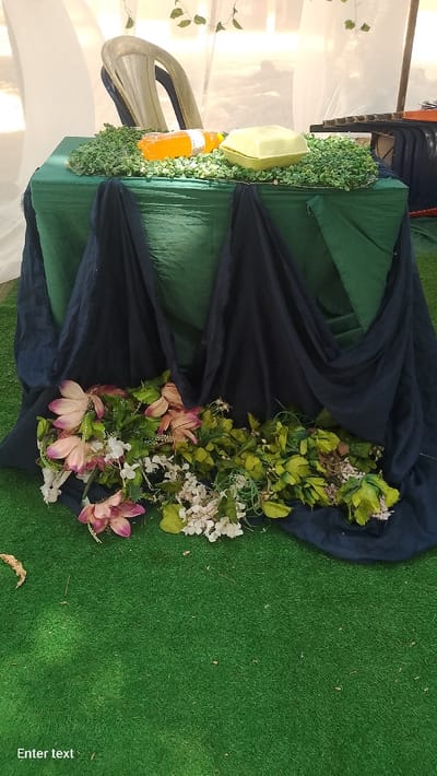 An event setup is depicted, presumably in Rinze, Nigeria. A rectangular table, covered with a dark green tablecloth, stands centrally. Dark blue fabric is draped vertically down the front of the table. The tabletop is adorned with a layer of small, artificial green foliage. Resting on this foliage are a clear plastic bottle containing an orange liquid with a white label, and a closed light-yellow foam food container. Behind the table, a light-colored plastic chair is partially visible against a background of white translucent drapes, which appear to be part of a temporary structure or tent. Artificial green leaves are sparsely attached to these drapes. The floor is covered with artificial green turf. At the base of the table, on the turf, a collection of artificial flowers and foliage is arranged, featuring pink/purple large petals, small white flower clusters, and various green leaves. No individuals are present in the frame, and no actions or interactions are occurring.