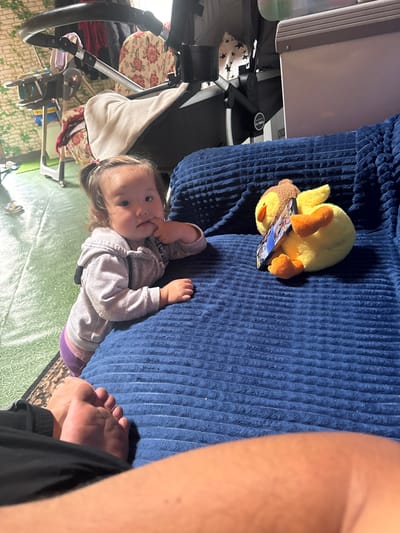 The image shows a baby lying on a blue textured surface, possibly a sofa or bed, with a stuffed yellow duck toy placed near her. The duck is holding a smartphone. To the left of the baby, a part of a baby stroller is visible, along with some patterned cushions. The baby is looking towards the camera with her finger in her mouth. Her attire consists of a light-colored hooded shirt and purple leggings. In the foreground, the legs and feet of an adult are visible, wearing black pants. The scene appears to be indoors, with a green mat or carpet on the floor. The lighting suggests it could be daytime, although the overall lighting is somewhat dim. No specific weather conditions are discernible. The context provided indicates the location is Yabogan, Russia.