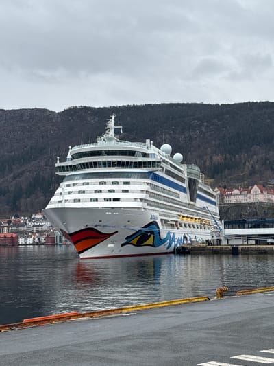 A horizontal shot of a large cruise ship docked in Bergen, Norway. The ship, with a distinctive painted bow featuring a large eye design, dominates the scene. It has multiple decks lined with windows and balconies. In the background, a mountain range looms over the city, and a cluster of buildings is visible along the waterfront. The water appears calm. A paved area with markings runs along the right side of the image, suggesting a dock or pier. The sky is overcast, creating a muted and slightly gray tone to the overall scene.