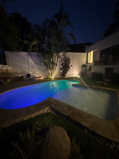 At Tres de Mayo, Mexico, the scene depicts an illuminated outdoor area at night, featuring a swimming pool with multicolored lighting that transitions from blue to green and white. The pool has a water feature, possibly a fountain, spraying water into the pool.
Adjacent to the pool, there's a patio with an inflatable alligator, a black chair, and toys. The area is bordered by a white building with lit windows and a surrounding landscape of trees, bushes, and grass. The overall scene is a mix of a recreational area integrated with natural elements.