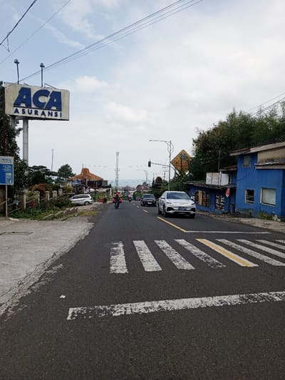 The image displays a daytime street scene in Ciloto, Indonesia, under a partly cloudy sky.
In the foreground, an asphalt road features a faded white and yellow pedestrian crossing with parallel white lines. A concrete sidewalk borders the left side of the road.
On the left, a tall sign structure prominently displays "ACA ASURANSI" in large blue lettering on a white background, positioned above a smaller blue sign partially obscured, but showing "PLN" and "IP". Behind the signs, a stone fence encloses an area with vegetation, leading to a building with a multi-tiered brown roof. A silver minivan is parked on the shoulder near this building, and several individuals are visible in its vicinity.
On the right side of the road, a two-story building with a vibrant blue facade and brown trim stands adjacent to the street. Its ground floor appears to house commercial establishments. Beyond this building, dense green foliage is present. A street light with a traffic signal is visible further down the road.
Traffic consists of several vehicles: a silver sedan (license plate L 1573 LZF) is positioned in the right lane, facing the viewer. Behind it, a dark-colored sedan proceeds in the same direction. In the opposing lane, a motorcyclist on a pink-colored scooter travels away from the viewer. Additional vehicles are discernible in the distance.
The background presents a hazy panorama of distant hills or mountains under the cloudy sky, with a telecommunication tower visible on the left horizon.