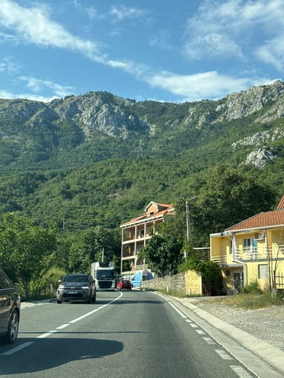 A view of a road in Košljun, Montenegro, with a mountainside covered in dense green trees in the background. The road curves to the right, with two lanes marked by white lines. Several vehicles are on the road, including a dark SUV in the foreground, a white truck, and a red car. Two buildings, one orange-roofed and multistory, and another yellow with a red-tiled roof, are visible on the right side of the road. The sky is blue with scattered white clouds.