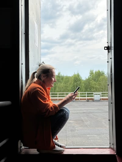 A blonde woman with her hair in a ponytail, wearing an orange fleece jacket and blue jeans, is squatting inside a doorway, looking at her smartphone. She is positioned on the left side of the frame, with the open doorway revealing a view of a grey, paved rooftop area, a white railing, and a dense forest of green trees under a cloudy sky. The doorway is framed by dark, metallic walls. The location is identified as being in the city of Tashtagol, Russia. The woman is the sole human subject in the image. Her action is focused on her phone. The environment appears to be an outdoor urban or industrial setting with natural elements visible in the background.