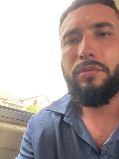 The image is a close-up, angled shot of a man's face and upper torso. He has dark hair, a beard, and is wearing a blue collared shirt with a repeating pattern of "CG" logos. In the background, out of focus, are the roofs of several houses. The bright, washed-out sky suggests it is daytime. The location is identified as Novy Khushet, Russia.