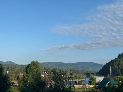 The image depicts a rural landscape with a river flowing through a valley, bordered by forested hills and mountains. The sky is blue with scattered white clouds. In the foreground, there are several houses with red and green roofs, interspersed with trees and utility poles. The scene is bathed in soft, natural light, suggesting it was taken during the daytime, possibly early morning or late afternoon. The location is identified as Kebezen, Russia.
