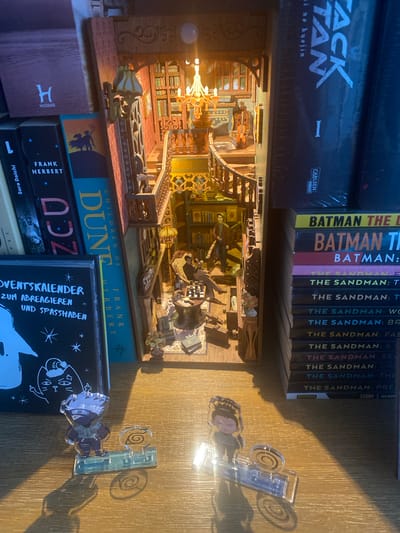 The image displays a detailed miniature library diorama placed on a wooden shelf. The diorama features a multi-level interior with intricate architectural details, including a grand staircase, railings, lamps, and bookshelves filled with tiny books. Several miniature figures are depicted within the diorama, engaged in activities like reading and sitting in chairs.
To the left of the diorama, there are several books. One book has a black cover with white text that reads "ADVENTSKALENDER ZUM ABREAGIEREN UND SPASS HABEN". Next to it are more books, including one with a teal cover and text that appears to be "DUNE CHILDREN OF FRANK HERBERT". Further to the left are books with dark covers and white text, including "FRANK HERBERT" and what looks like "Sora Daichi".
To the right of the diorama, a stack of books with the title "THE SANDMAN" is visible, along with some "BATMAN" books.
In the foreground, two small anime-style acrylic figurines stand on the wooden shelf. The figurine on the left depicts a character with spiky white hair and a mask. The figurine on the right shows a character with dark hair tied up. Both figures are standing on clear acrylic bases with a swirl design.
The environment appears to be an indoor setting, likely a room with bookshelves. The lighting suggests a warm, ambient glow, possibly from the diorama's internal lights and external ambient light filtering from the left.
Based on the available visual information, there is no specific information within the image that can confirm its location in Hildesheim, Germany.