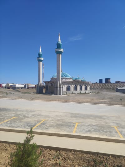 The image depicts a modern mosque with two tall minarets, a central dome, and several smaller domes. The mosque is constructed with light-colored stone and features turquoise accents on the domes and minaret tops. In the foreground, there is a paved area with yellow parking lines and a low concrete barrier separating it from a patch of dry earth with sparse vegetation. In the background, a line of white and red trucks is visible, suggesting a nearby industrial or construction area. The sky is a clear, bright blue, indicating fair weather. The location is identified as the city of Egizbulak, Uzbekistan.