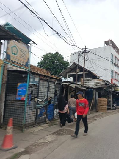 This image depicts a street scene in Dayeuhkolot, Indonesia, under an overcast sky. In the immediate left foreground, an orange traffic cone rests on the edge of a paved road. The left side of the street is lined with a series of weathered, single-story structures with corrugated metal roofs. The first structure features a sign with a hexagonal logo and text, and a blue-and-white poster is affixed below it. Further along this side, another structure exhibits similar construction and a blue tarp-like material.
On the right side of the street, a similar row of structures continues, including one with stacked, circular woven cages. In the background, a multi-story white building with visible red trim stands prominently. A wooden utility pole with multiple cross-arms and numerous dense bundles of electrical and communication wires is positioned in front of this white building. Overhead, a complex network of black utility lines crisscrosses the frame against the light gray sky. A large, dark green tree is visible between the structures in the midground and the taller white building.
Three individuals are present on the paved street and sidewalk. In the right foreground, a man walks towards the right, wearing a red long-sleeved shirt, dark pants, a blue baseball cap, and sandals. A light-colored object is slung over his right shoulder. Behind him, a woman, facing away from the viewer, wears a black head covering, a patterned top, and dark pants, carrying a dark bag in her left hand. Further behind her, a third person, in a light brown top and dark shorts, also walks away from the viewer. The paved road appears slightly damp in areas.