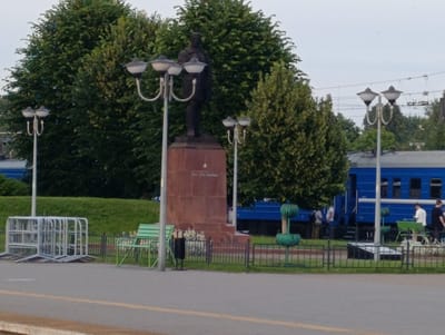 The media file depicts an outdoor scene at what appears to be a public space, possibly near a train station in Orsha, Belarus. The central subject is a tall, dark statue of a man standing on a reddish-brown, rectangular pedestal. The statue and pedestal are flanked by ornate, multi-globed lampposts. In the background, a bright blue train is visible, suggesting a transportation hub. The setting includes lush green trees and grass, indicating a temperate climate, likely daytime with overcast skies.
In front of the statue and pedestal, there's a black metal fence enclosing a grassy area. Within this fenced area, a green metal bench and a trash receptacle are visible. To the left of the statue, a stack of silver metal barriers or barricades are present, suggesting a temporary setup or a storage area. Further to the right, a small, decorative green fountain or water feature stands near the fence.
Several people are present in the scene, though most are indistinct. On the right side of the image, near the blue train, a few individuals in casual attire can be seen. One person in a white shirt is standing, while another in a dark shirt is also visible. Other figures are more subtly present, either near the train or within the general vicinity of the monument.
The overall mood of the image is calm and ordinary, capturing a typical day in this location. There is some visible text on the pedestal of the statue, though it is somewhat obscured and difficult to read clearly from the image quality. The presence of the train, monument, and public amenities points towards this being a public area within Orsha.
