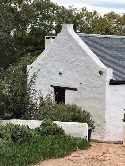 A close-up, canted-angle shot shows a white-walled, rural building with a gray corrugated metal roof. A small, barred window is visible on the wall, partially obscured by leafy bushes. A section of white wall extends towards the viewer, covered in greenery. A sandy path runs alongside the greenery. The location is Theewaterskloof Nu, South Africa.