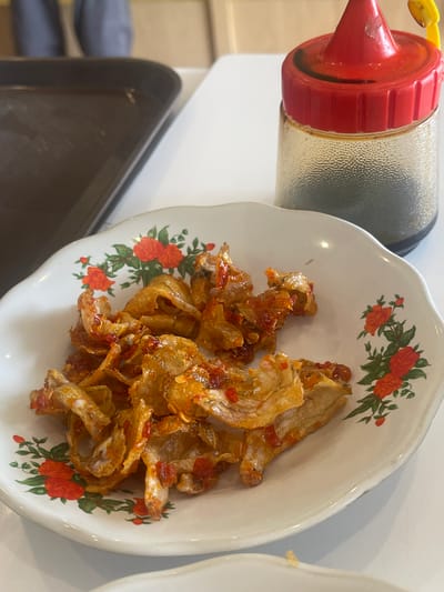 In Medan, Indonesia, a plate of fried anchovies in a chili sauce is presented on a white table. The plate is decorated with a floral pattern of red roses and green leaves. Next to the plate is a condiment bottle with a red cap, containing a dark liquid, likely soy sauce or a similar condiment. In the background, a dark brown tray is visible, and a person's legs are out of focus. The lighting is natural, suggesting an indoor setting like a restaurant or home.