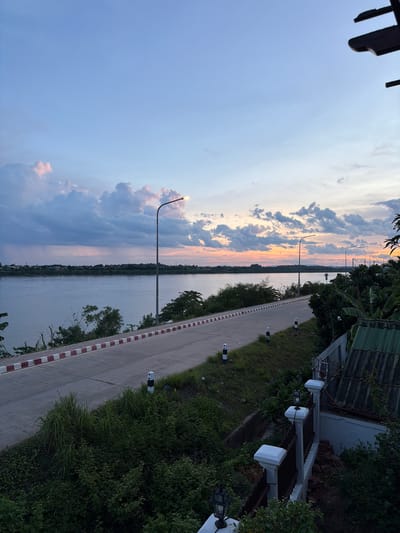This is a photograph taken at dusk along the Mekong River in Vientiane Capital, Laos. The sky displays a gradient of blue, with pink and orange hues from the setting sun visible near the horizon. Clouds are scattered across the sky, with some illuminated by the last rays of sunlight. A wide, paved road runs along the riverbank, bordered by a white and red striped curb. Several lampposts are spaced along the road. Dense green vegetation, including trees and bushes, lines the side of the road, and some of it extends down towards the river. In the foreground, to the right, is a portion of a house with a green corrugated roof and a white fence with decorative posts and lamps. The river is calm and reflects the colors of the sky. The scene depicts a tranquil evening by the river in Vientiane.