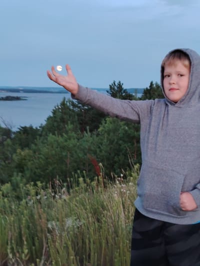 A young boy, wearing a grey hoodie and dark patterned shorts, stands in a grassy, overgrown area with trees and a body of water in the background. The sky is a pale blue, suggesting dusk or dawn. The boy has his left arm extended, and his palm is facing upwards, with a small, bright object, possibly the moon, resting on it. The scene appears to be outdoors, in a natural setting.