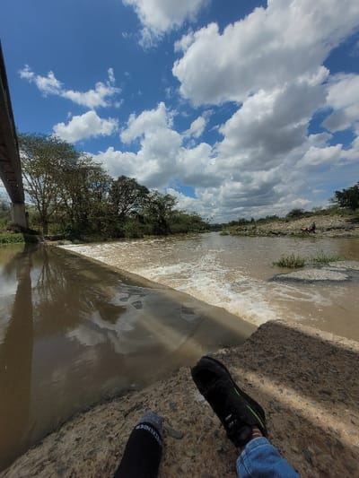 The image displays a river scene within Kinanie ward, Kenya, captured from a low perspective. The sky is bright blue, featuring numerous large white cumulus clouds. In the immediate foreground, the lower legs and feet of an individual are visible, resting on a rough, textured concrete surface. The left foot wears a black sock with white "WEEKENDER" lettering, while the right foot is clad in a black athletic shoe with green accents and blue denim jeans. Directly beyond the foreground, a concrete weir extends across a brown, murky river, creating a turbulent, foamy cascade of water downstream. Upstream, the river appears calmer. A concrete bridge structure, likely an overpass, is partially visible spanning the river on the far left. Both riverbanks are densely lined with green trees and shrubbery. On the far right bank, two indistinct human figures are visible near the water's edge, appearing to be seated or resting.