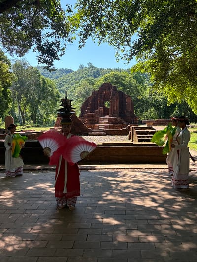 The image depicts a cultural performance at My Son Sanctuary, a UNESCO World Heritage Site located in the Quang Nam province, near the city of Đà Nẵng, Vietnam. In the foreground, four individuals are dressed in traditional Vietnamese attire. The central figure wears a vibrant red dress and holds two large red fans. To the left, another individual in a white and red outfit holds a green fan. On the right, two more individuals in similar white and red attire stand together. They are positioned on a brick-paved walkway, with dappled sunlight casting shadows from the surrounding trees. In the background, the ancient brick ruins of My Son Sanctuary rise, nestled amidst lush green trees and hills under a clear blue sky. The atmosphere suggests a sunny day and a serene historical setting.