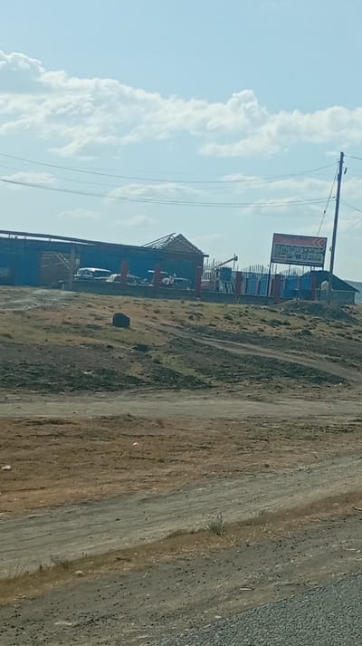 A building with an orange fence and a business sign stands on a slight rise. A few cars are parked inside the fenced area. The roof of the building is partly damaged. A large rock sits in the dry field in front of the building. The foreground is dominated by a dusty, gravel-covered road. The sky is light blue with scattered white clouds. Power lines stretch across the top of the frame.