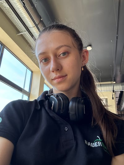A young woman with brown hair and light-colored eyes is pictured in a close-up, tilted selfie. She is wearing a black polo shirt with a green logo on the left chest, which reads "BAUER". Over-ear Marshall headphones are resting on her head. Her hair is pulled back and falls over her shoulder. She is looking directly at the camera with a neutral expression.
The background shows industrial-style architecture with exposed pipes and lighting fixtures, along with large windows that suggest an indoor setting, possibly an office or a modern workspace. The natural light coming from the windows indicates it is daytime. The overall impression is casual and personal. The text on her shirt is clearly visible.