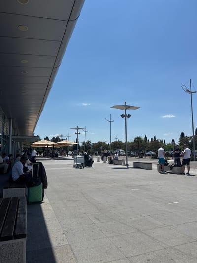 The image shows an outdoor area outside an airport terminal on a sunny day. Several people are visible, some sitting on benches and others standing. There is luggage present, including a green suitcase. The architecture of the terminal building is modern, with a large overhang and circular lights visible on its underside. In the background, there are trees, a parking lot with cars, and streetlights. The ground is paved with large gray tiles. The sky is clear and blue. The scene appears to be at Podgorica Airport, located in Golubovci, Montenegro.