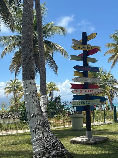 The image shows a directional signpost with colorful arrow-shaped signs pointing to various locations, including "Rio de Janeiro," "Martinique 162 km," "La Desirade 50 km," and "Les Saintes 40 km." The signpost is located in a tropical environment with palm trees. The sky is blue with some clouds. The image is likely taken in or near the city of Le Gosier, France, considering the distances listed on the signpost and the tropical environment typical of Guadeloupe. The ground is covered with grass. The image is taken from a low angle.