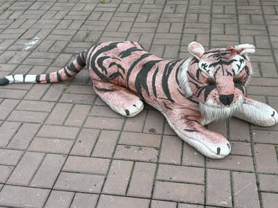 A large, plush tiger stuffed animal lies on a brick-paved ground. The tiger is predominantly pinkish-brown with black stripes, mimicking a real tiger's pattern. It has white fur around its face and chest, and its eyes are round and dark. Its tail extends to the left and ends with white and black rings. The tiger appears to be lying down, with its paws splayed out on the ground. The brick paving is laid in a basket-weave pattern, and small patches of grass are visible in the gaps between the bricks. There are some light-colored smudges on the bricks, possibly chalk or paint. The scene appears to be outdoors, as indicated by the natural light and the presence of grass. There is no visible text in the image. The overall impression is of a toy left outside, possibly a whimsical or slightly melancholic sight. The time of day and weather are not explicitly indicated, but the lighting suggests it is daytime with overcast or diffused sunlight. The location context provided is Каменка, Russia.