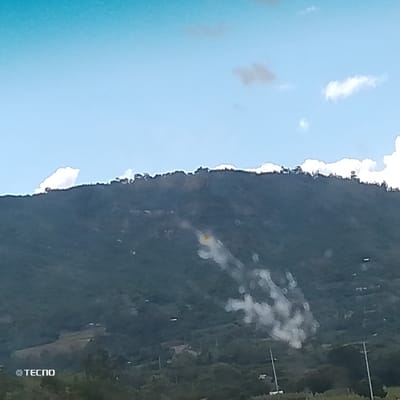 The scene depicts a large, heavily vegetated mountain or hill located in Kola ward, Kenya, under a bright blue sky with scattered white cumulus clouds. A significant plume of white smoke is observed originating from the mid-slope on the right side of the mountain, extending diagonally downwards. Immediately above and slightly to the left of the main smoke plume, a distinct yellow patch, consistent with a flame or burning material, is visible. The mountain features dense green foliage and subtle lighter patches on its lower slopes, potentially indicative of terraced land or clearings. The foreground reveals two dark utility poles with cross-arms on the right, suggesting the presence of power lines or similar infrastructure. Small, indistinct structures are also visible on the lower hillside. No individuals are discernible within the frame. A "© TECNO" watermark is present in the bottom left corner.