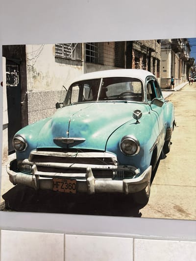 The image displays a two-tone vintage automobile, predominantly light blue with a white roof, showing significant wear, dents, and paint degradation. The vehicle features chrome bumpers and grille. An orange license plate, located centrally on the front bumper, displays "CUBA" above the alphanumeric code "HFZ360." This evidence indicates the scene is not situated in West Felton, United Kingdom. The car is parked on a light-colored, possibly unpaved or heavily worn street, angled slightly towards the right. To the immediate left is a dilapidated building with aged, cracked plaster and a dark recessed entrance or doorway, alongside windows with metal bars. The street extends into the background, flanked by more old, multi-story buildings, some featuring barred windows or architectural details. Several indistinct human figures are visible walking in the distance on the right side of the street, with one individual in a blue shirt standing closer on the right sidewalk. The scene is illuminated by bright daylight, casting clear shadows. No direct actions or interactions are occurring in the immediate foreground.