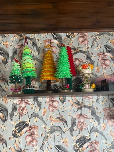 The image displays a decorative arrangement, possibly a shelf or rack, mounted against a wall adorned with a floral wallpaper pattern. The wallpaper features a light blue or gray background with brown, beige, and gray floral and leaf motifs. A wooden door or panel is visible on the left side of the frame.
The decorative arrangement includes several miniature Christmas-style trees constructed from yarn or similar soft material. The trees vary in color, including green, red, and yellow/orange. They are decorated with small ornaments, beads, or decorative accents. Additionally, there are what appear to be small floral arrangements using flowers in shades of yellow, orange, and white. Pinecones can also be seen as part of the decorations.
The specific location as Gorun, Bulgaria is not identifiable from the image alone. There are no people, subjects, actions, or interactions visible in the scene.