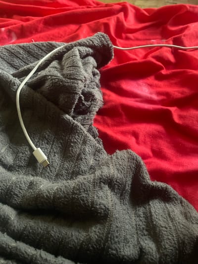 The image shows a close-up of a crumpled gray fleece blanket with a white USB-C cable resting on it, partially on a bright red fabric background. The blanket has a textured, fuzzy surface with thin, vertical stripes. The red fabric is creased and appears to be a sheet or similar material. The scene is indoors, with the lighting suggesting an ambient setting. There are no people visible in the image. The overall impression is one of casual disarray. The image location in Линёвка, Russia cannot be determined from the visual information provided.