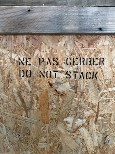 A close-up image depicts a section of a shipping container or panel in Aulnay-sous-Bois, France. The primary subject is an Oriented Strand Board (OSB) panel, characterized by its surface composed of compressed, irregularly oriented wood strands in various shades of light brown and tan. Above the OSB panel, a horizontal, weathered wooden plank, exhibiting a grey-brown hue and visible grain, spans the upper portion of the frame. Two dark circular indentations, consistent with nail or screw heads, are present in the plank. Stenciled in black capital letters across the OSB panel are the phrases "NE PAS GERBER" and, directly beneath it, "DO NOT STACK". No individuals are visible. The scene appears to be an interior or sheltered environment, typical of a warehouse, storage facility, or industrial setting.