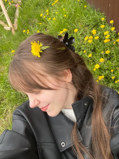 A woman lies in a field of dandelions in Karmaskaly, Russia. The woman, who appears to be Caucasian, lies on her back, facing the camera. She wears a black leather jacket and a white sweater. Her long, brown hair is accessorized with a black flower clip and a single dandelion. The ground surrounding her is covered in green grass and numerous yellow dandelions. A wooden fence is partially visible in the background, indicating the edge of the field. The lighting is bright, suggesting it is daytime.