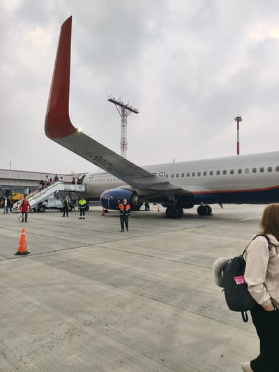 The image depicts an outdoor scene at Emelyanovo Airport, Russia, on an overcast day. In the foreground, a woman with long brown hair is walking away from the camera, carrying a black backpack with a neck pillow and a pink item protruding from it. She is wearing a light-colored jacket and dark pants.
The main subject of the image is a large airplane, likely an Aeroflot aircraft, as indicated by the visible text on its fuselage. The plane is parked on the tarmac, and a set of mobile stairs is attached to its left side, with passengers disembarking. Several individuals in uniform, possibly ground crew, are visible around the stairs and the plane, some directing passengers and others performing duties. One person in an orange and black high-visibility vest is standing near the plane's nose wheel, looking upwards.
Several orange traffic cones are placed on the tarmac, marking areas or guiding movement. In the background, a tall radio mast with lights is visible against the cloudy sky. The overall environment is an airport tarmac, characterized by concrete pavement and large aircraft.