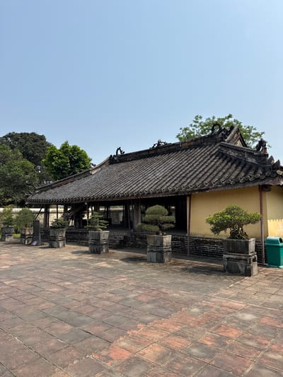 The image depicts a traditional Vietnamese building with a tiled roof in Kim Long Ward, Vietnam. The building features intricate architectural details, including ornate roof ornaments and supporting pillars. The tiled roof is composed of individual tiles arranged in a pattern. A tan building is in the background, with a green trash can on its right side. Trees surround the structure, adding greenery to the scene. The foreground consists of a paved walkway made of rectangular tiles. The sky is blue and clear.