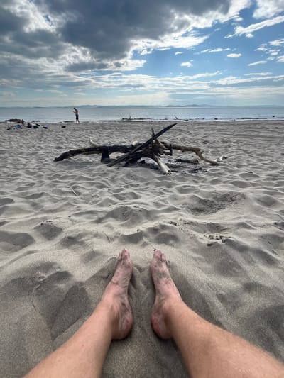 This image depicts a beach scene with a foreground dominated by sandy ground and a person's bare legs and feet. In the midground, there is a collection of driftwood, some of which appears to have been burned, suggesting a campfire site. Further in the background, the sea stretches out to the horizon, with hills visible in the distance under a cloudy sky with patches of blue. A few people are scattered on the beach, with one person standing near the water's edge. The scene is set on a beach, likely in the city of Krasnoturansk, Russia, given the context provided.