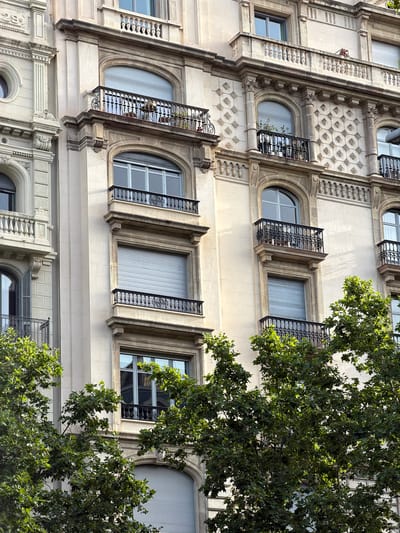 This is a close-up, eye-level, outdoor, daytime, vertical, eye-level shot of the facade of an ornate, pale stone building in Barcelona, Spain. The building features multiple floors with arched windows, some of which have black wrought-iron balconies. The windows are covered by light gray roller shutters, and some balconies have potted plants. The building's architecture includes decorative columns, cornices, and textured stone panels. In the foreground, dense green foliage from trees partially obscures the lower portion of the building, with sunlight filtering through the leaves. The scene is tranquil and architectural, highlighting the building's details and the surrounding greenery.