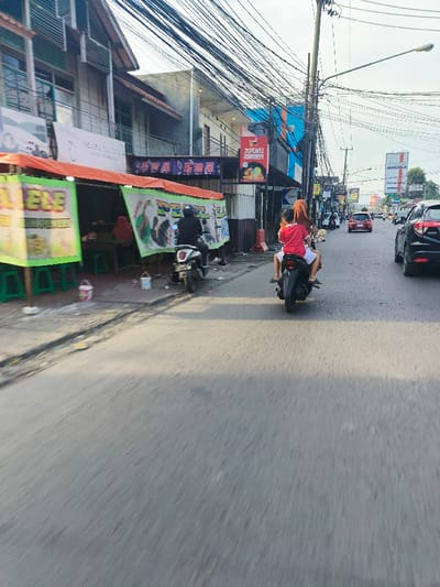 The image shows a street scene in Bojongsoang, Indonesia, at the coordinates -6.974 latitude and 107.636 longitude. The street is paved and lined with buildings on both sides. There are numerous power lines strung overhead. A motorcycle with two people on it is driving down the center of the street. The driver is a woman wearing a brown headscarf and the passenger is a young boy wearing a red shirt. Other vehicles, including cars and a motorcycle, are also visible on the street. To the left of the street is a storefront with a green awning and green and yellow signage. There are several trash cans and a few people standing nearby. The buildings in the background are mostly one or two stories tall and constructed with concrete. There are trees and other vegetation visible in the background. The sun is shining, and the sky is blue. The image appears to be taken from a moving vehicle looking out the window.