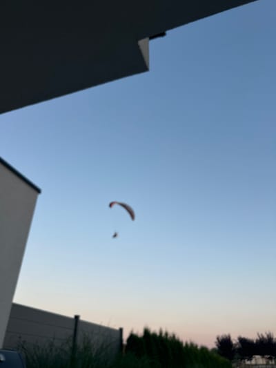 The image, taken in Warzymice, Poland, depicts a paraglider soaring through a pale blue sky, likely during dawn or dusk. The paraglider, a dark shape with red accents, is suspended by lines from a canopy. Below the paraglider, a small human figure is visible. To the left of the frame, the corner of a modern building with light-colored walls and a dark overhang is present. In the foreground, a dark fence runs horizontally across the lower part of the image, with dark green foliage and trees visible behind it. The overall scene is slightly blurred, suggesting motion or a soft focus effect.
