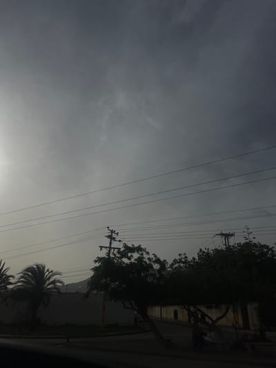 The image depicts a cloudy sky over a street scene in San Antonio, Venezuela. In the foreground, silhouetted palm trees and other foliage are visible along with utility poles and overhead wires. A wall runs along the background, behind which a distant hill is faintly discernible. Two individuals are present, one standing and another seated on a bench. The lighting suggests it is either early morning or late afternoon, with overcast conditions contributing to a muted atmosphere.