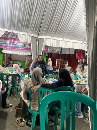 The image depicts an indoor gathering, likely a banquet or a community event, held under a white tent structure. The event appears to be taking place in Wedarijaksa, Indonesia, as indicated by the signage visible in the background. Several people, mostly women wearing hijabs, are seated around tables, engaged in conversation or observing the surroundings. Some individuals are standing, interacting with others or managing the event. Food and drinks are present on the tables, suggesting a meal is being served. The environment is functional, with plastic chairs and tables arranged for the gathering. The overall atmosphere seems informal and social.