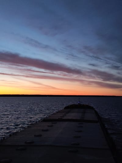 The image shows a view from the deck of a vessel at dusk. The deck is gray and features numerous evenly spaced, round hatches. The dark water stretches out to meet the horizon, where a dark treeline is visible. Above the horizon, the sky displays a gradient of colors from orange to pink to deep blue. Clouds are present in horizontal bands. A small light is visible at the end of the deck.