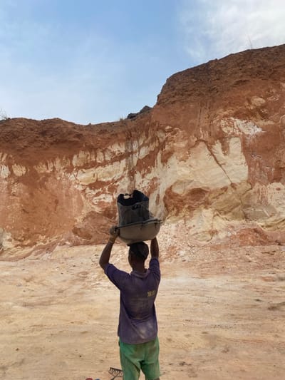 The scene depicts a male individual observed from behind, standing in what appears to be an excavation or quarry site in Asaga Amangwu, Nigeria. He is wearing a purple short-sleeved shirt, green shorts, and a dark head covering, possibly a cap. The individual is carrying a large, dark-colored bucket or container nested within a wider, metallic basin, both balanced on his head. His arms are raised, hands positioned to steady the load. The ground directly beneath and around him is flat, dusty, and reddish-brown. In the lower left foreground, a metal rake and several other small, indistinct objects are visible on the ground. The background is dominated by a substantial, exposed earth cliff face, displaying distinct stratification of reddish-brown topsoil and lighter, yellowish-white rock or clay layers. The cliff exhibits vertical striations and areas of erosion. Above the cliff, a pale blue sky with minimal cloud cover is visible.