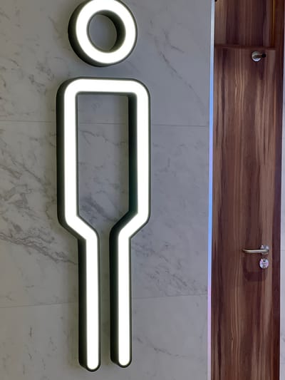 A modern, illuminated restroom sign for men is mounted on a marble-tiled wall next to a wooden door with a silver handle. The sign is a stylized, outlined figure of a person with a circular head and a rectangular body that tapers to two legs. The outline is illuminated with bright white light. The background wall is made of large marble tiles with a grey veined pattern. To the right of the sign, a wooden door is visible, featuring a natural wood grain texture and a silver door handle and lock. A thin, vertical strip of iridescent light runs between the wall and the door. The image is taken from a slightly low angle, looking up at the sign and door. The overall setting suggests a clean and contemporary public restroom. The image does not provide specific information about the city of Derry/Londonderry, United Kingdom.