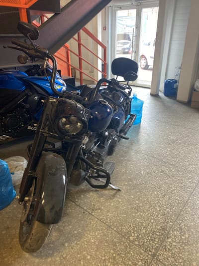 The image depicts a close-up view of several motorcycles parked indoors, potentially a storage area or garage, in the city of Svkh Voronezhskiy, Russia. The motorcycles are predominantly blue and black. Other details:
* The setting appears to be a commercial or industrial building with a tiled floor.
* There is a metal staircase with orange railings visible on the left side of the frame.
* A window is visible in the background, revealing a view of a parked car.
* Several blue plastic bags are scattered around the motorcycles.