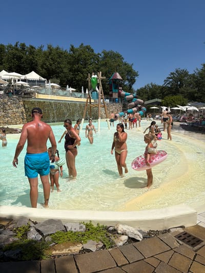A group of people are enjoying a sunny day at a water park. The scene is a shallow pool with clear, bright water, with a sandy or light-colored bottom. Several individuals are standing or wading in the water. In the foreground, a man wearing blue swim trunks and a black baseball cap is facing away from the camera, his back to the viewer. To his left, a young girl in a swimsuit and a striped shirt stands with a pink inflatable ring patterned with red hearts. Further into the pool, other people are visible, some in swimsuits, appearing relaxed and enjoying the water.
Behind the pool, a large water slide structure with colorful segmented tubes is visible. Beyond that, lush green trees form a backdrop under a clear blue sky. There are also white umbrellas and lounge chairs set up, suggesting a recreational area. The image is captured in daylight, likely midday, given the brightness and the shadows cast. The overall atmosphere appears to be one of leisure and fun, characteristic of a summer day at a water park. There is no visible text in the image. The location context provided is Cavriglia, Italy, which aligns with the warm, sunny weather and outdoor recreational setting.