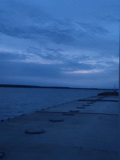 The image depicts a waterfront view at dusk or dawn. The sky is predominantly blue with cloud cover. Below the sky, a dark treeline or distant landmass stretches across the horizon. A body of water fills the middle ground, reflecting the dim light from the sky. In the foreground, a concrete pier or walkway extends into the water. There are several recessed circular shapes evenly spaced on the surface of the pier. A structure with a blue light is visible on the pier in the distance. There are no discernible people or interactions captured in the scene. Based on location information this is likely the Volga River waterfront area of Topornya, Russia.